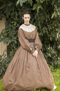 Danielle Yrulegui dressed in an 1860's Civil War day dress with a hoopskirt and petticoats. She's facing the camera with hands folded in front of her.