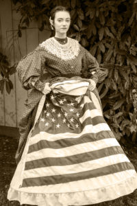 Danielle Yrulegui dressed in an 1860's Civil War day dress with a hoopskirt and petticoats. She's lifted her dress and petticoats to show a Union flag beneath, looking at the camera. The picture is sepia-toned.