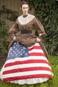 Danielle Yrulegui dressed in an 1860's Civil War day dress with a hoopskirt and petticoats. She's lifted her dress and petticoats to show a Union flag beneath, looking towards someone out of frame..