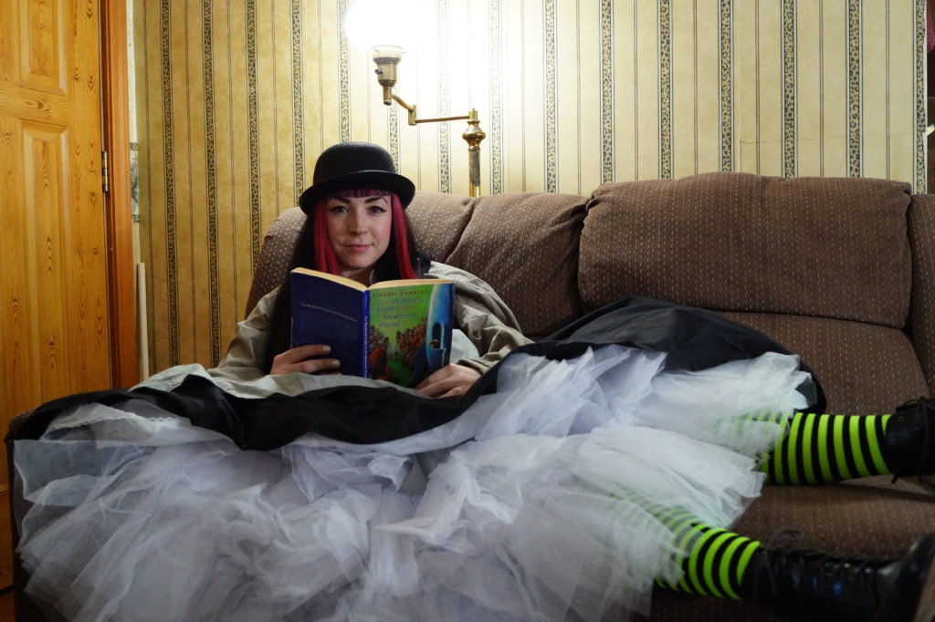 A steampunk woman in pink hair and a bowler hat on a couch, skirts spread out all over the place and showing crinoline tulle, along with neon green striped stocking. She's reading the book The Wonderful Flight to the Mushroom Planet, while looking at the viewer